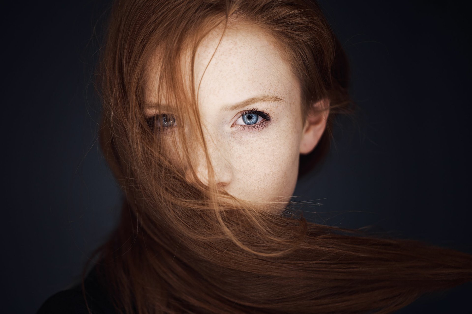 Close-up of a redheaded female model with blue eyes and freckles, her hair partially covering her face against a dark background.