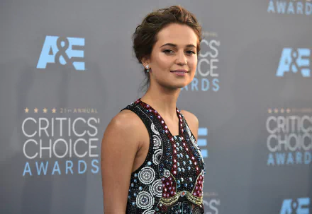 Alicia Vikander, the Swedish actress, smiles confidently with her brown eyes, showcasing her unique style at the Critics' Choice Awards against a branded backdrop.