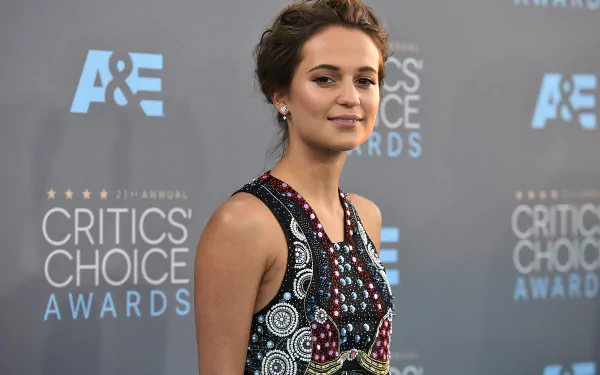 Alicia Vikander, the Swedish actress, smiles confidently with her brown eyes, showcasing her unique style at the Critics' Choice Awards against a branded backdrop.