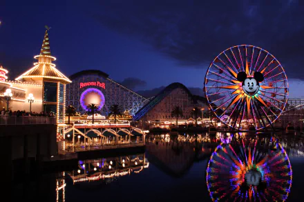 A vibrant night scene at Disneyland California features a colorful ferris wheel, roller coaster, and sparkling lights reflected on the water, capturing the magic of an amusement park.