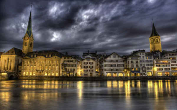 Dusk in Zurich, Switzerland: dramatic clouds and glowing lights reflect on the river, historic houses and church towers line the man-made waterfront. HD PC desktop wallpaper.
