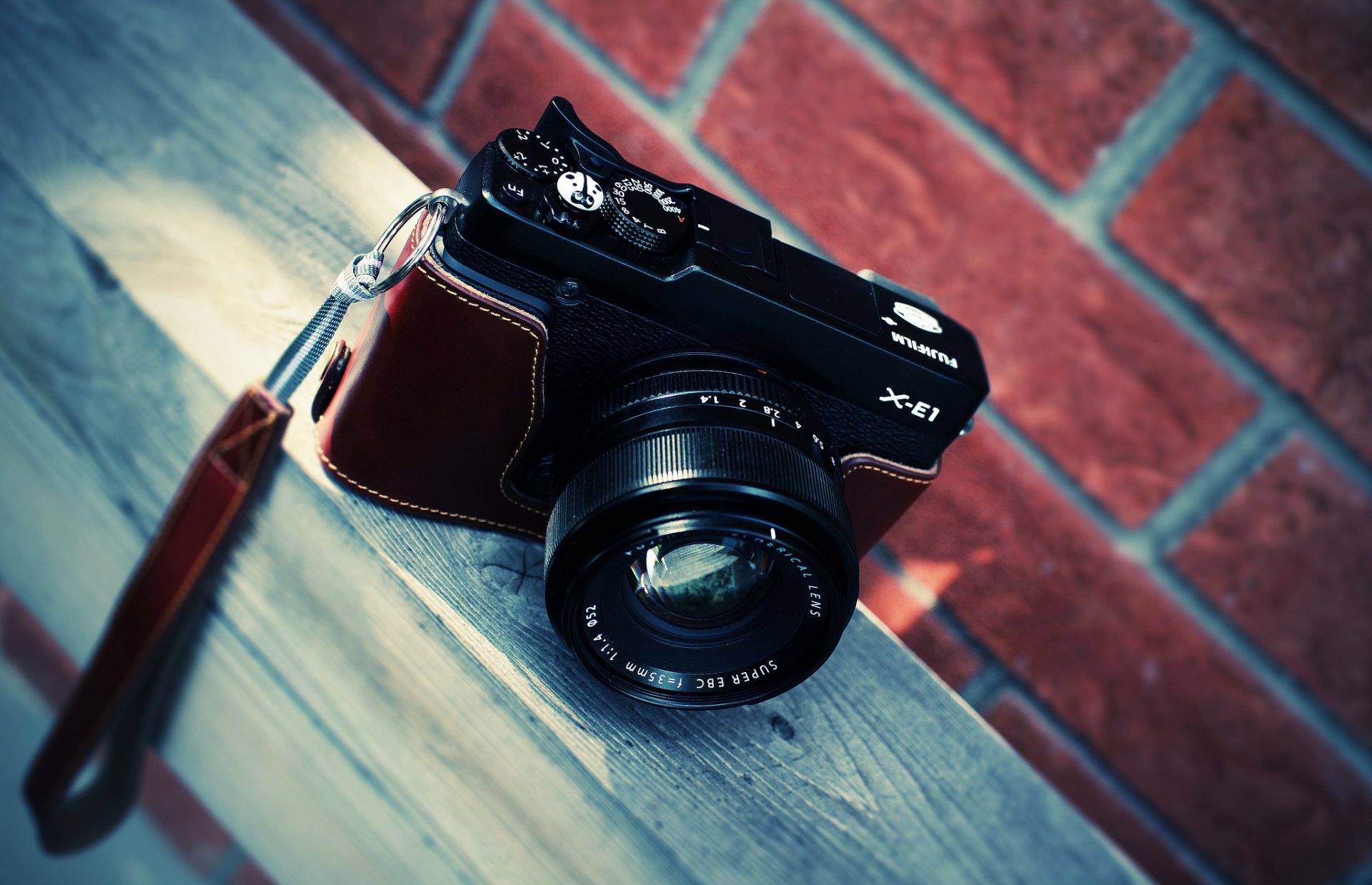 HD desktop wallpaper featuring a man-made camera with a leather case resting on a wooden surface against a brick wall background.