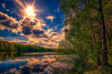 HD PC desktop wallpaper/background: sunlit river bordered by trees, dramatic clouds and bright sun in the sky reflected in calm water — vivid nature scene with clear tree reflection.
