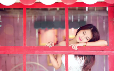 A brunette Asian woman with lipstick leans against a red window frame, eyes closed in a peaceful mood. This high-definition wallpaper captures a serene and contemplative moment.