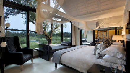 A serene bedroom featuring a large bed, plush pillows, a cozy chair, and a picturesque window view of a lush landscape, all under a soft canopy, creating a tranquil safari-inspired atmosphere.