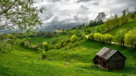 A vibrant HD desktop wallpaper of a lush green landscape featuring a rustic barn, rolling grass-covered hills, mountain backdrop, and a distant village under a cloudy sky.