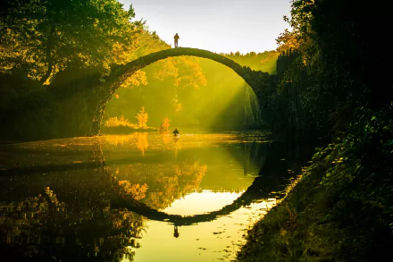 A serene view of the Devil's Bridge in Germany, reflecting on a calm river. The golden hues of nature surround the man-made structure, creating a tranquil atmosphere.