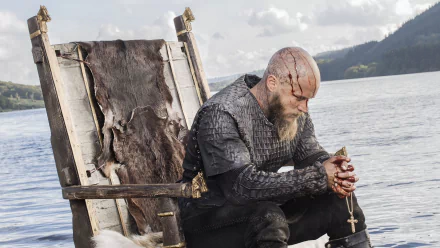 Ragnar Lothbrok sits pensively on a throne by the water in this HD wallpaper from the TV show Vikings (2013), capturing a moment of reflection.