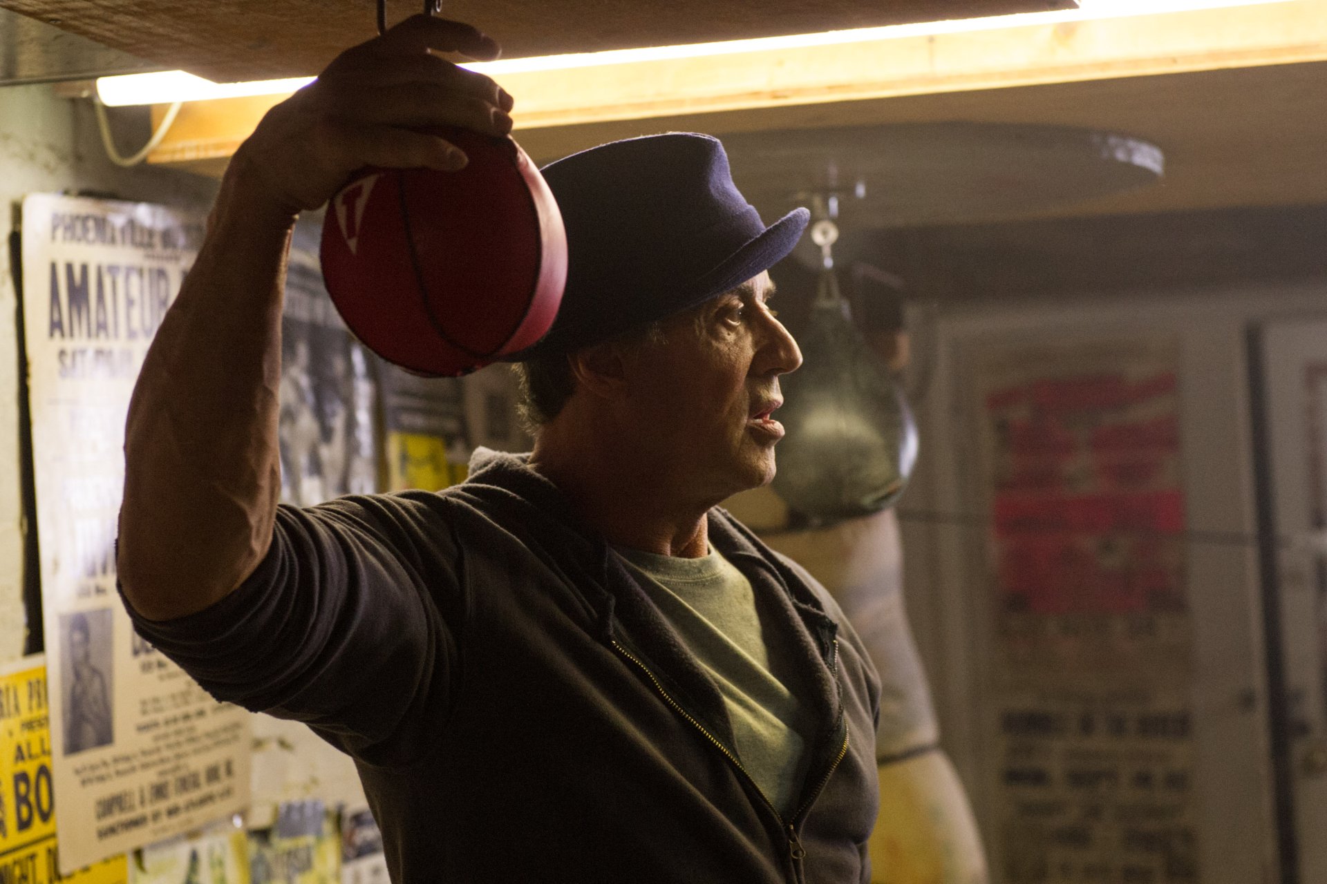 2K Quad HD desktop wallpaper: cinematic movie-style still of an older boxing trainer in a dim gym, cap on, holding a speed bag — evocative of the Rocky/Creed boxing films.