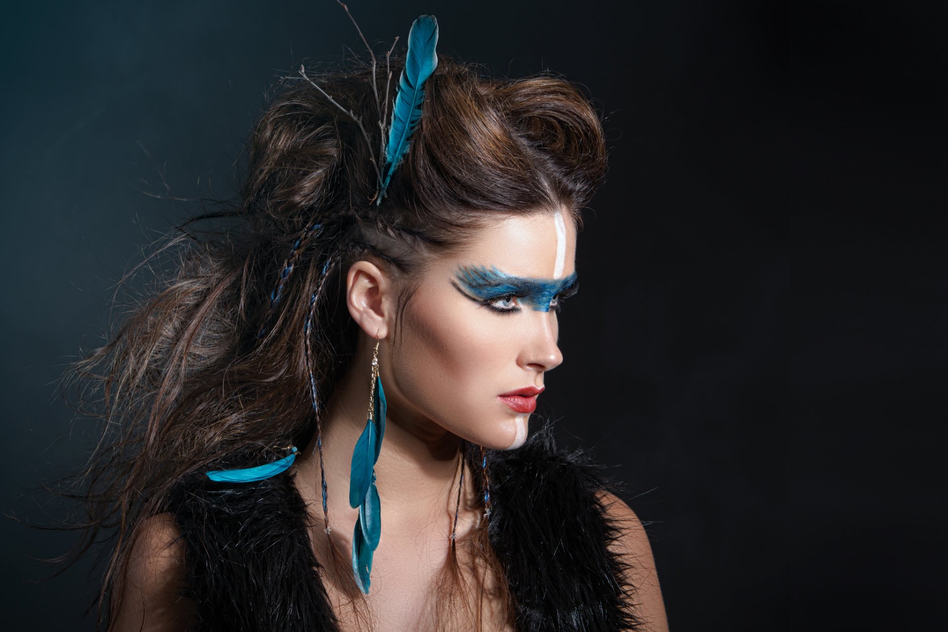 A striking brunette model showcases bold makeup with blue accents and dramatic feathers, capturing attention with her vibrant blue eyes against a dark background.