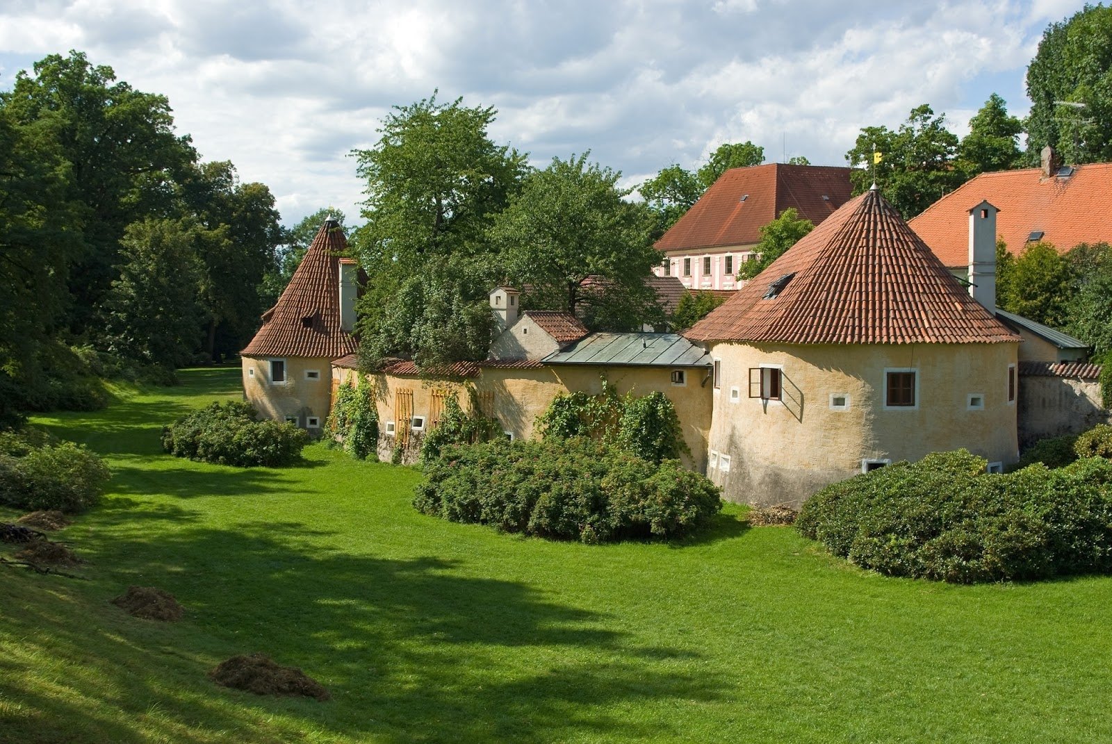 HD desktop wallpaper of an old Czech Republic village: man-made terracotta-roofed houses and stone walls amid green lawns and trees under a cloudy sky.