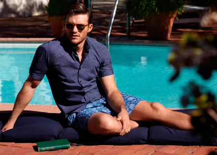Scott Eastwood, American actor and celebrity, relaxes by a pool wearing sunglasses and casual summer attire in this HD PC desktop wallpaper and background.