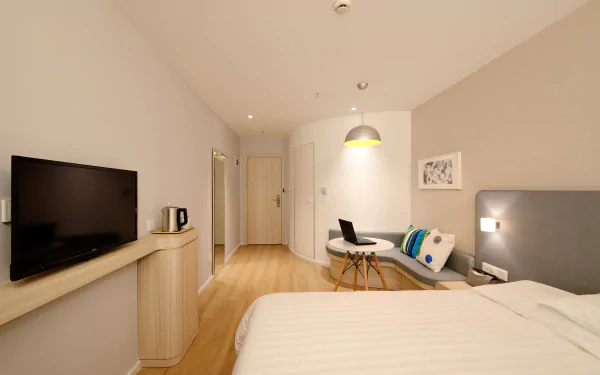 A modern hotel room with a bed, cushioned seating area, table with a laptop, wall-mounted television, and soft lighting in 4K Ultra HD resolution.