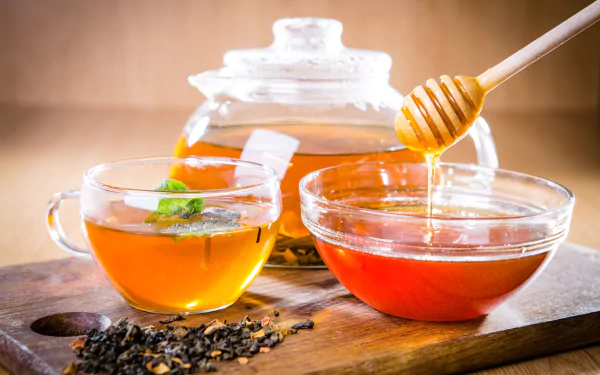 A 4K Ultra HD desktop wallpaper featuring a glass cup of tea, a bowl of honey with a honey dipper, loose tea leaves, and a glass teapot on a wooden surface.