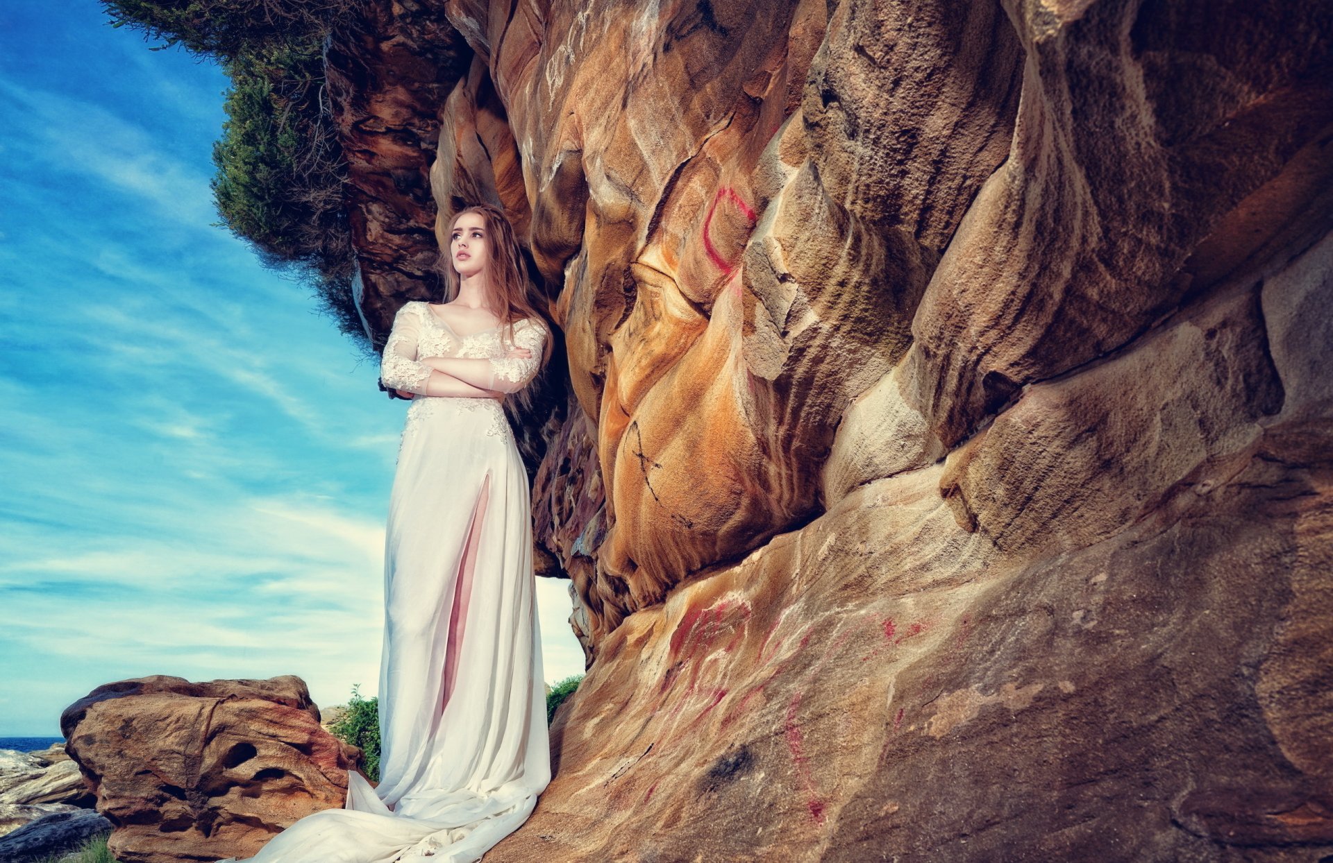 A brunette woman with brown eyes in a white dress stands against a rocky outdoor background, exuding a serene mood under a blue sky. HD desktop wallpaper.