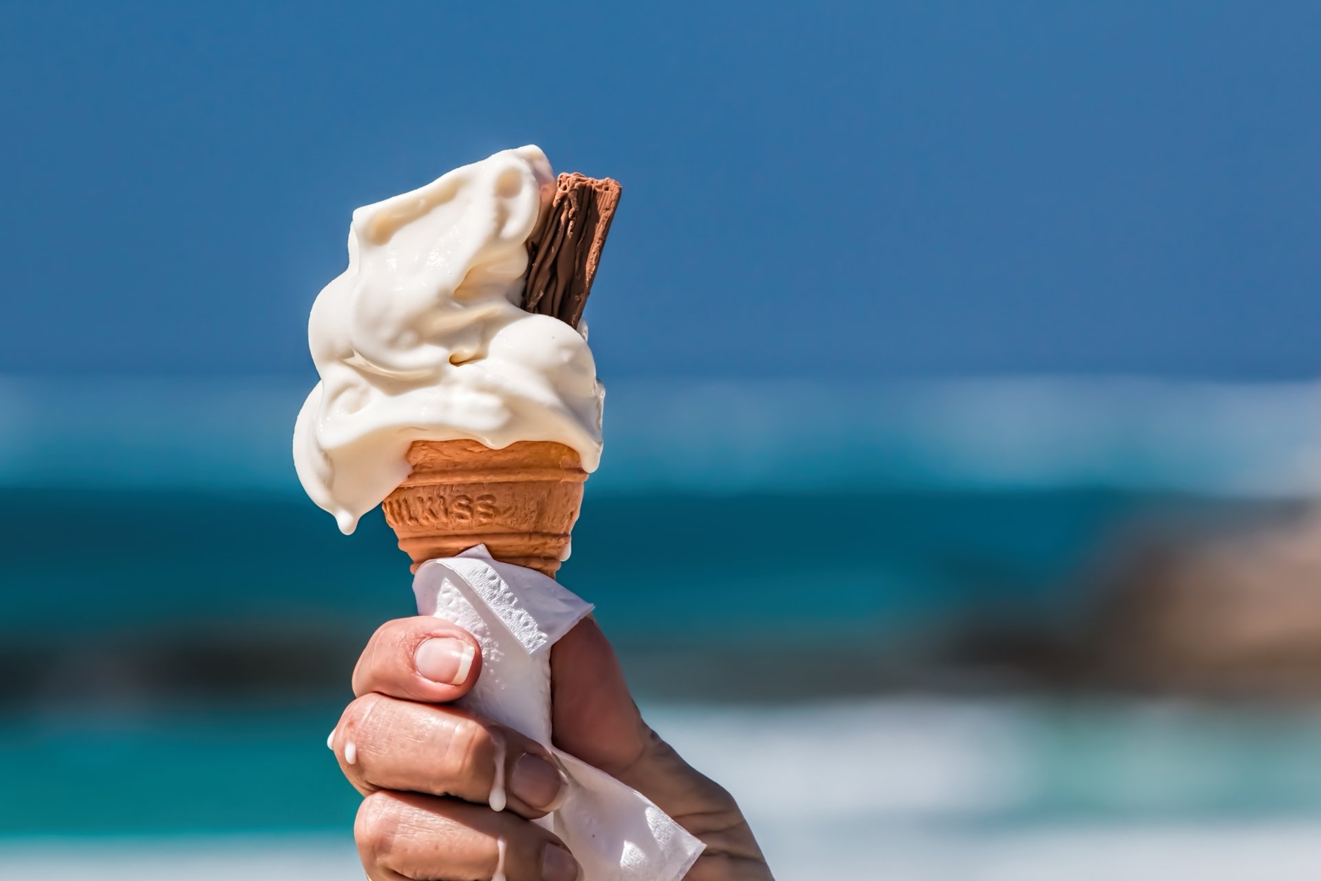 A hand holds a soft-serve ice cream cone with a chocolate stick against a blurred blue ocean background in a vibrant 4K Ultra HD dessert-themed wallpaper.