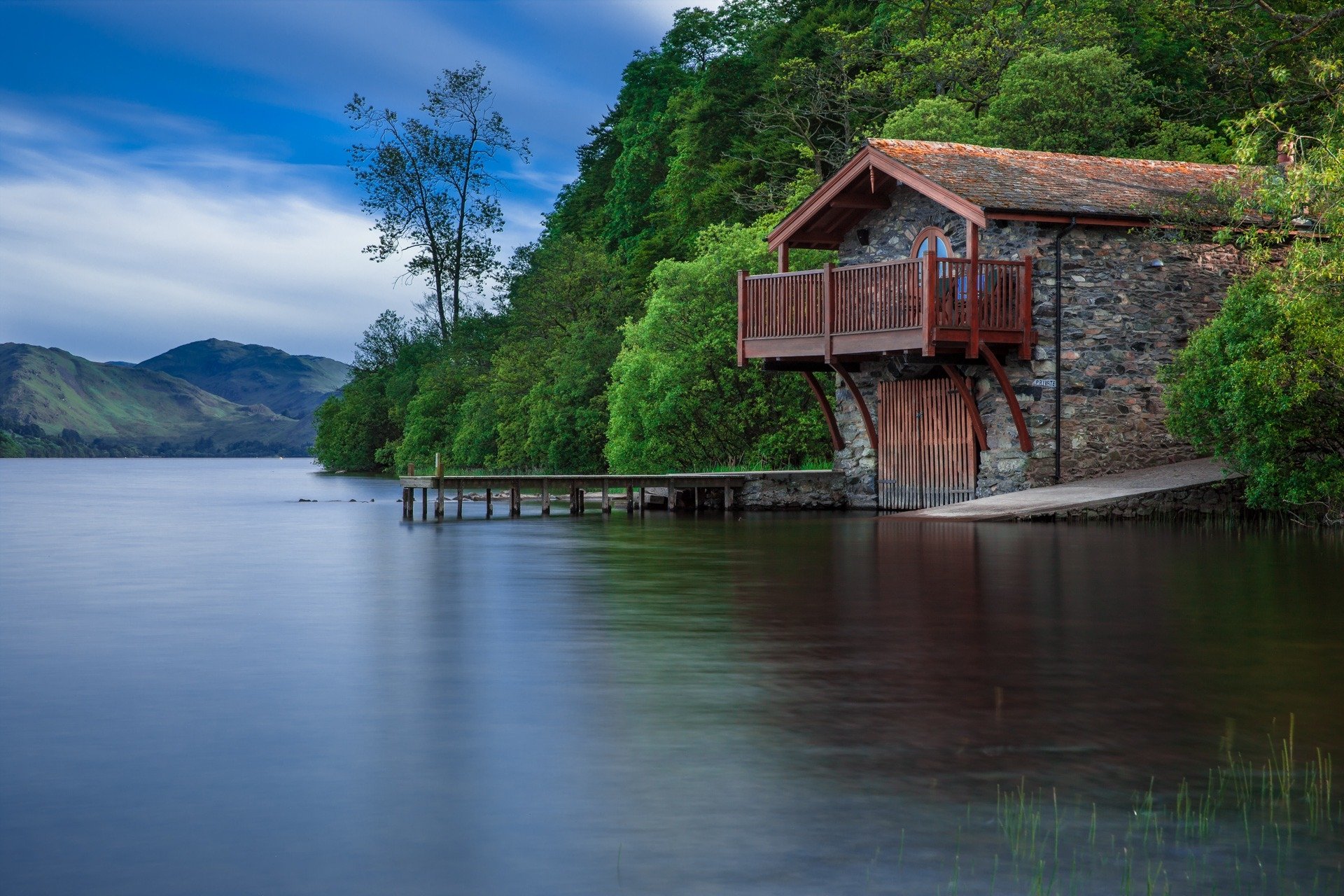 A serene HD desktop wallpaper of a brick boathouse by a lake, surrounded by lush trees and hills under a partly cloudy sky.