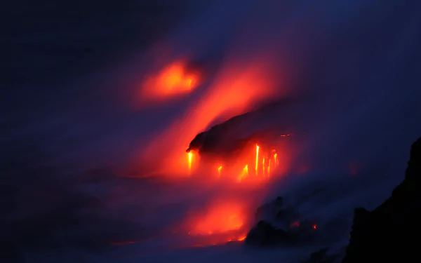5K Ultra HD PC desktop background: a night volcano in nature, glowing lava flows and smoking plumes casting red-orange light across dark coastal rocks.