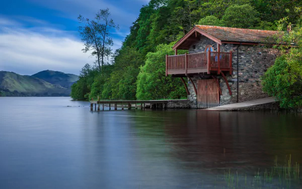A serene HD desktop wallpaper of a brick boathouse by a lake, surrounded by lush trees and hills under a partly cloudy sky.