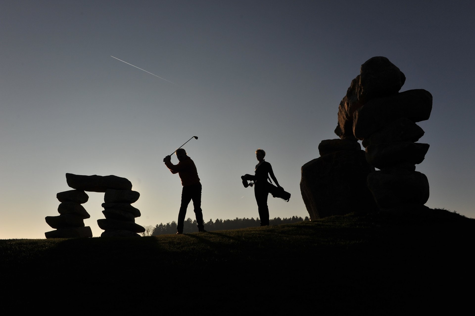 Silhouettes of a golfer swinging and another carrying a golf club at evening, set against a clear sky with stone formations, captured in 4K Ultra HD for a striking sports scene.