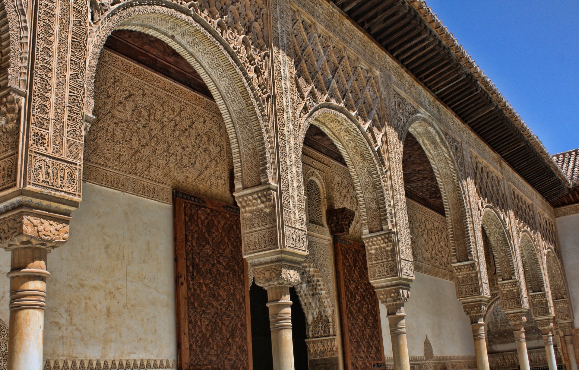4K Ultra HD desktop wallpaper of the Alhambra in Granada, Spain — carved Moorish arches, columns and wooden doors beneath a bright blue sky; man-made historic palace detail.