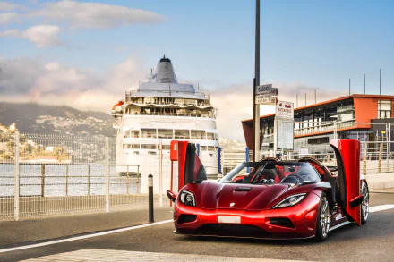 A striking Koenigsegg Agera R in vibrant red with its doors open, set against a backdrop of a cruise ship and modern waterfront architecture, creating a stunning HD desktop wallpaper.