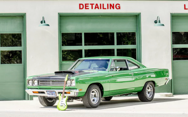 A vibrant green vintage Plymouth Road Runner muscle car parked in front of a detailing garage, accompanied by a retro electric guitar leaning against it.