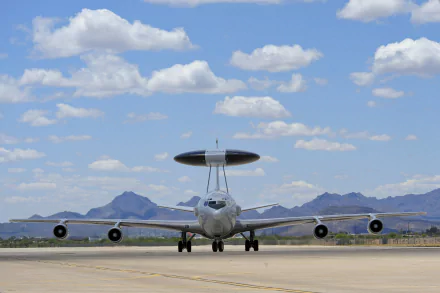 HD desktop wallpaper featuring a Boeing E-3 Sentry military warplane on a runway with mountains and a blue sky with scattered clouds in the background.
