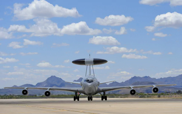 HD desktop wallpaper featuring a Boeing E-3 Sentry military warplane on a runway with mountains and a blue sky with scattered clouds in the background.