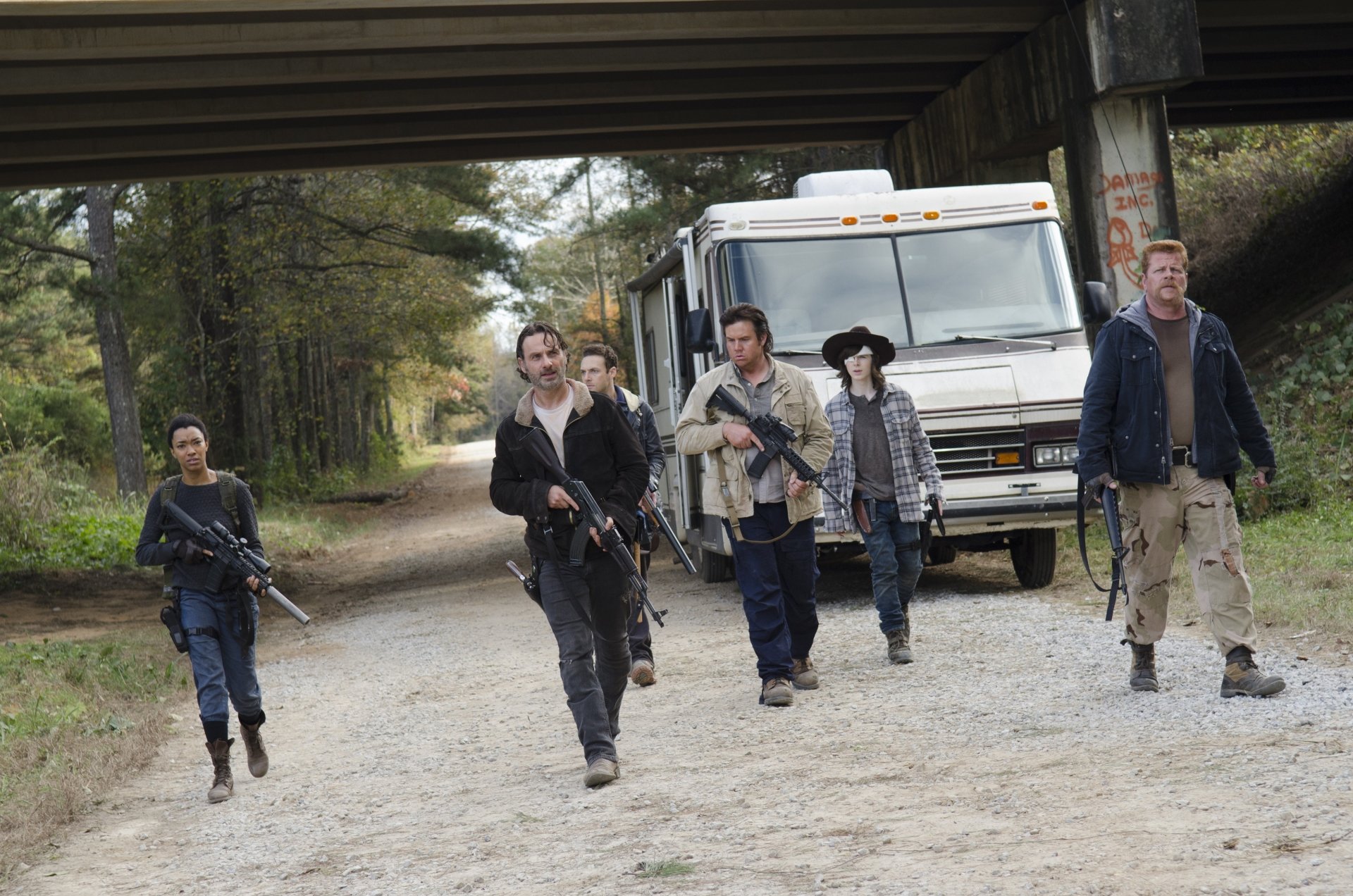 A scene from The Walking Dead featuring Rick Grimes and Carl Grimes alongside a group of survivors walking down a gravel road, with an RV parked in the background.