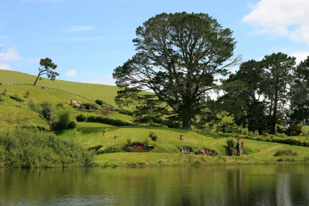 HD desktop wallpaper showcasing the man-made Hobbiton in Comarca, featuring lush green hills, large trees, and a reflective pond under a clear blue sky.