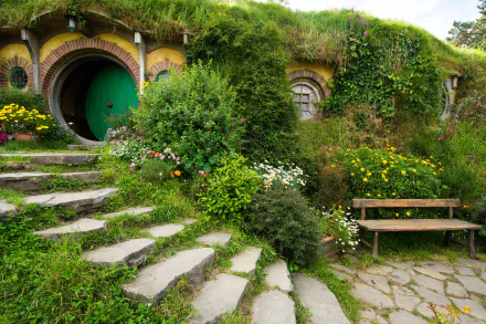 A 4K Ultra HD man-made Hobbiton scene featuring a grassy hillside with a round green door, stone steps, vibrant flowers, and a wooden bench in a lush garden setting.