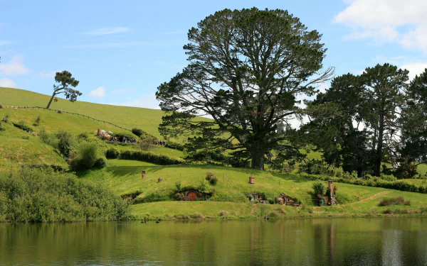 HD desktop wallpaper showcasing the man-made Hobbiton in Comarca, featuring lush green hills, large trees, and a reflective pond under a clear blue sky.