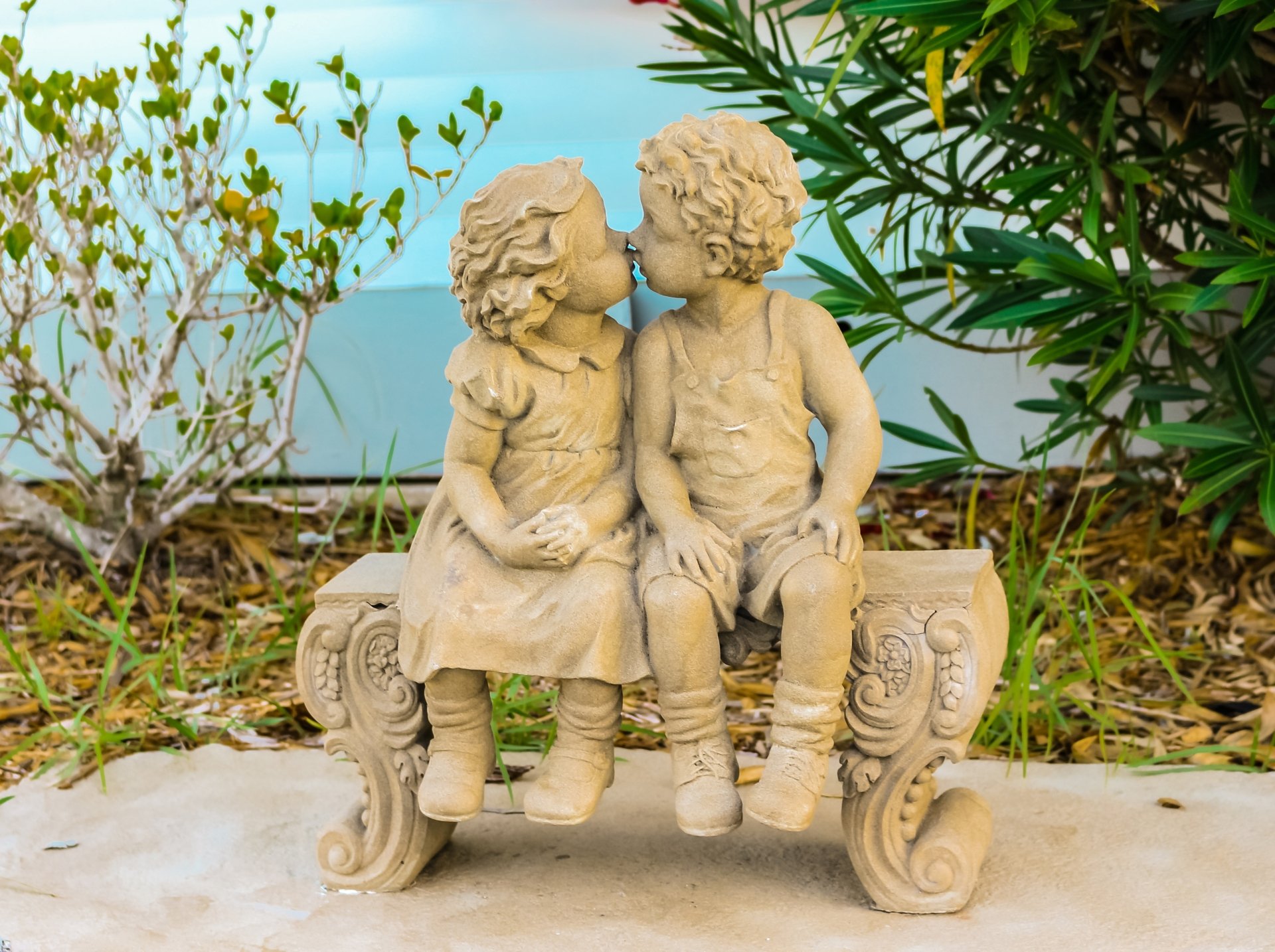 2K Quad HD PC desktop wallpaper showing a man-made stone sculpture statue of two children kissing on an ornate bench, set in a garden with surrounding foliage.