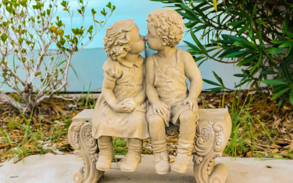 2K Quad HD PC desktop wallpaper showing a man-made stone sculpture statue of two children kissing on an ornate bench, set in a garden with surrounding foliage.
