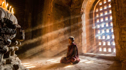 A serene little boy, dressed as a monk, meditates in a softly lit temple. Sunbeams filter through a patterned window, illuminating the peaceful atmosphere of this Buddhist setting.