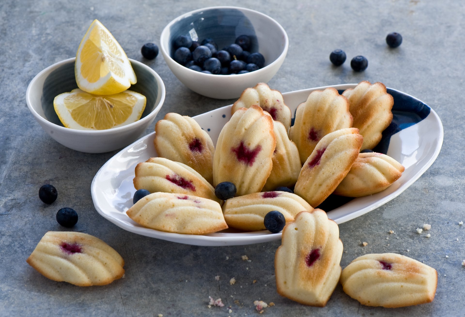 HD PC desktop wallpaper: blueberry madeleines on a white plate with scattered blueberries and lemon wedges on a gray tabletop.