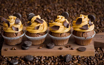 Four cupcakes topped with creamy swirls and chocolate pieces, surrounded by coffee beans and small cookies, set against a rich coffee bean background as an HD desktop wallpaper.