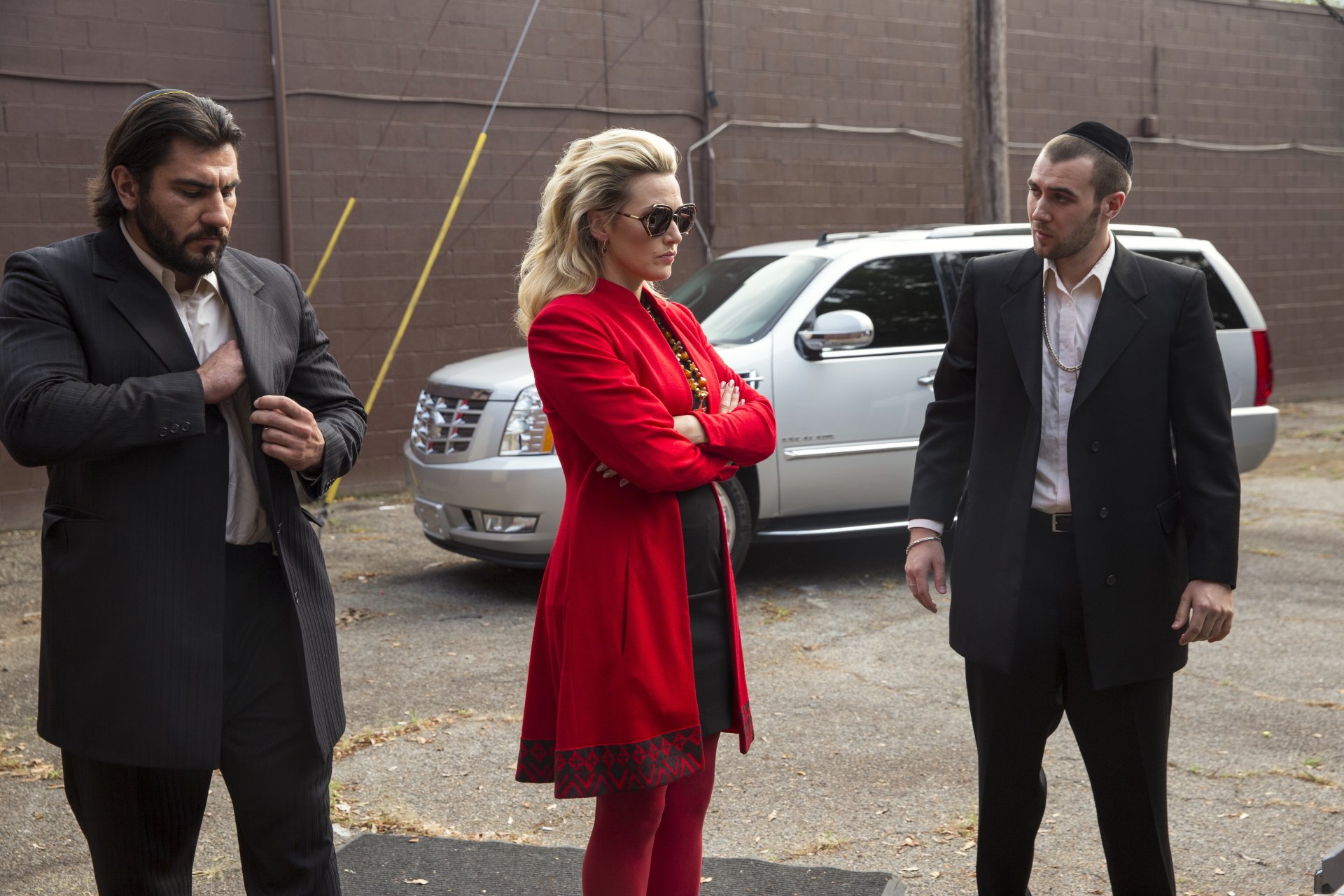 A scene from the movie Triple 9 featuring Kate Winslet in a striking red outfit, standing confidently between two men. The backdrop includes a sleek white vehicle. 4K Ultra HD wallpaper.
