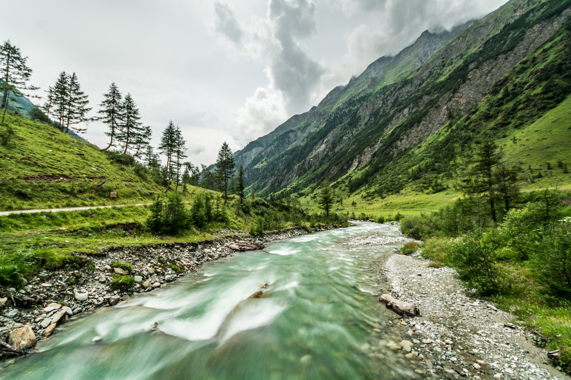 4K Ultra HD wallpaper of a lush green valley with a flowing river, surrounded by towering mountains under a cloudy sky, showcasing serene natural landscape beauty.