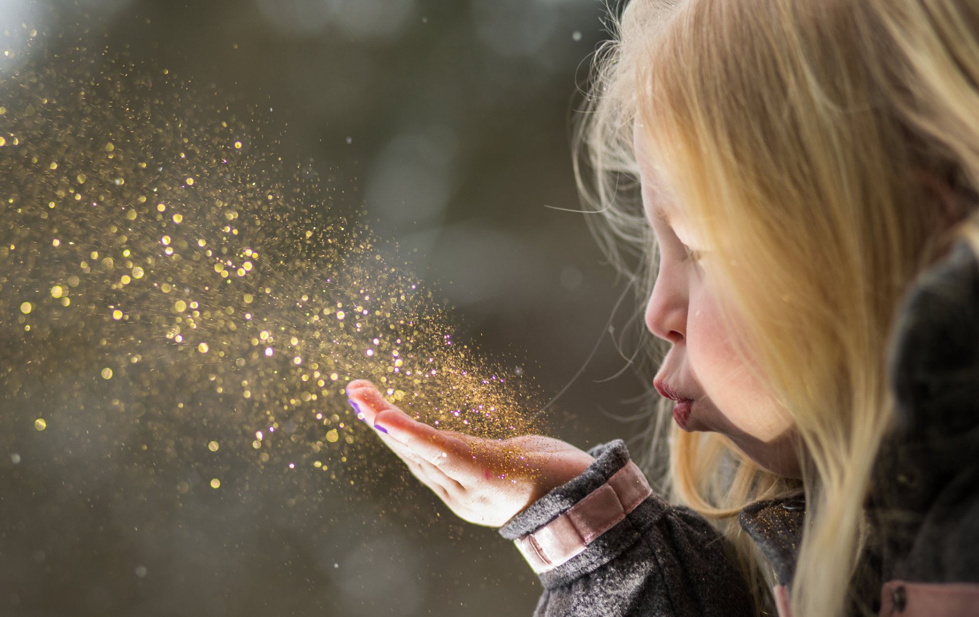 A blonde little girl blows shimmering glitter from her hand, captured in a detailed HD photography wallpaper background.
