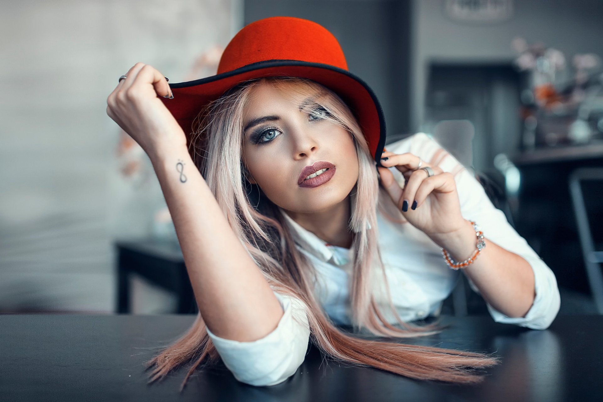 Blonde woman with blue eyes wearing a red hat, resting her head on a table in a stylish indoor setting, captured in HD for a desktop wallpaper background.