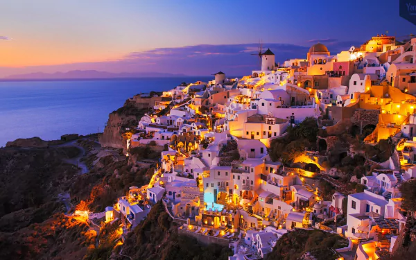 Santorini cliffside houses aglow at dusk, warm lights on white man-made buildings spilling toward the sea — HD PC desktop wallpaper/background.