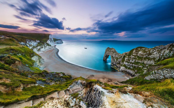 A stunning HD desktop wallpaper of England’s Dorset coastline featuring the iconic natural limestone arch of Durdle Door under a vibrant sky over the ocean horizon.