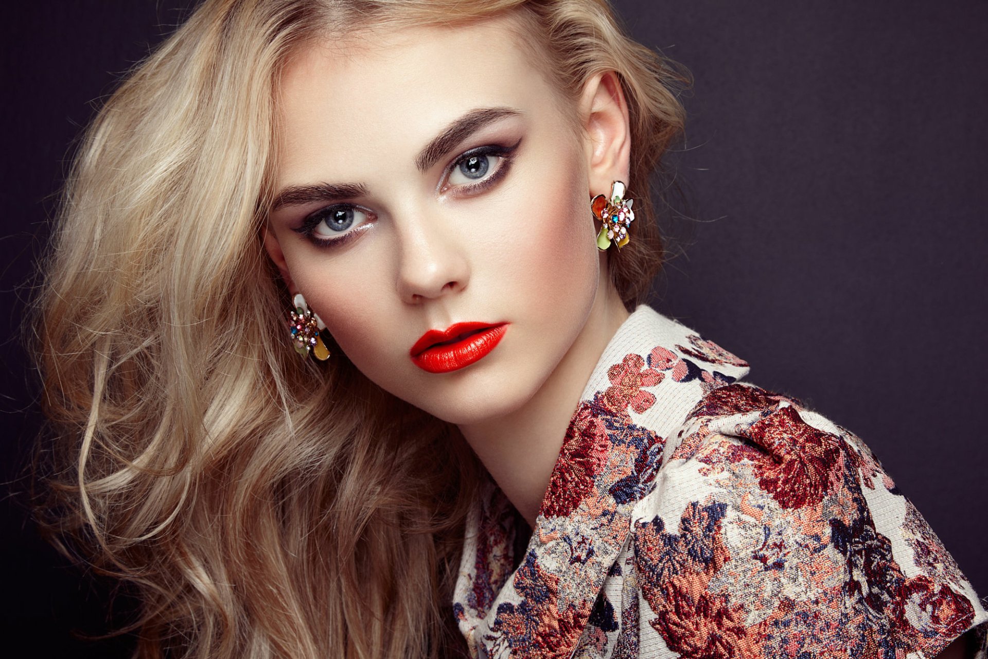 A stunning blonde model with striking blue eyes and bold red lipstick, adorned with vibrant earrings, poses elegantly against a dark background, showcasing beauty and fashion.