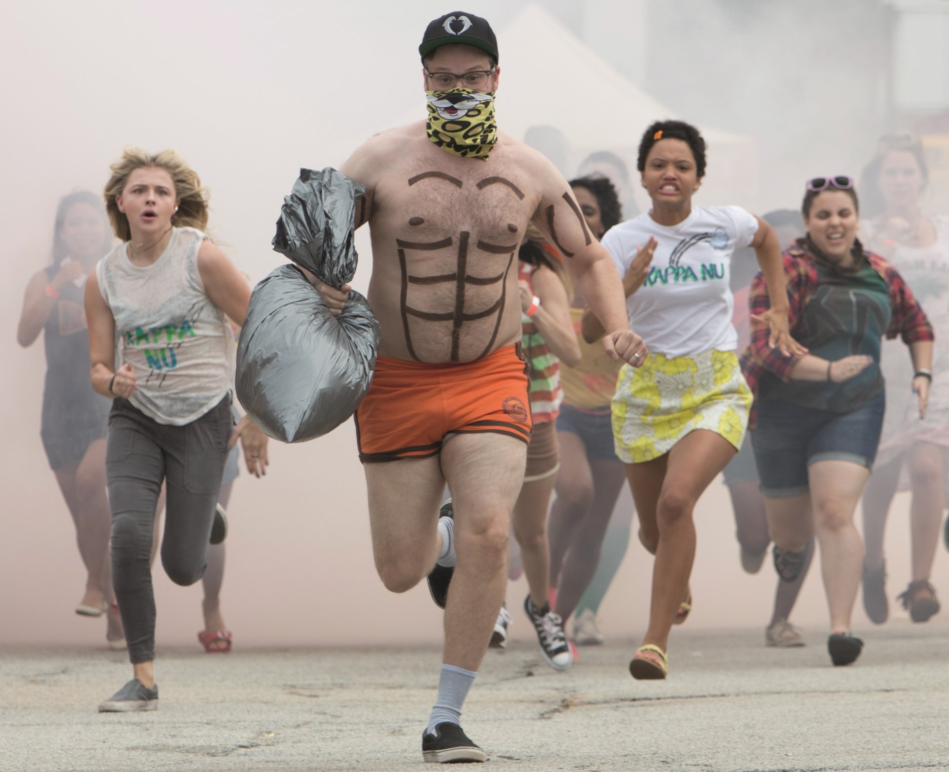 Group of women chasing a masked, painted-abs man in orange shorts through smoke — 2K Quad HD PC desktop wallpaper/background for the movie Neighbors 2: Sorority Rising.