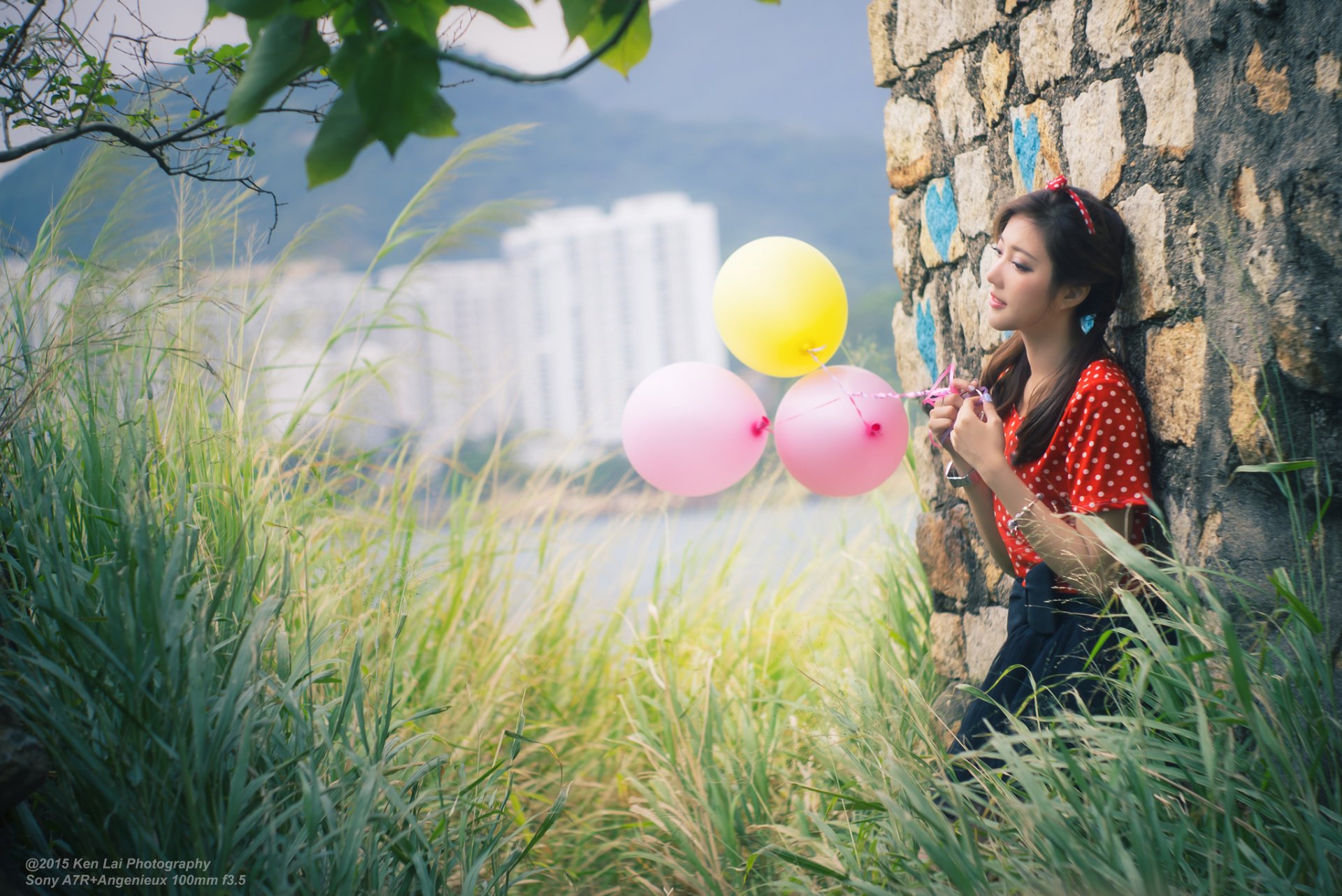 A woman named Wing Wing, holding colorful balloons, stands by a stone wall amidst tall grass with city buildings in the background. The HD wallpaper captures a serene outdoor moment.