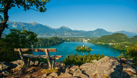 A serene landscape featuring a wooden bench overlooking Lake Bled, with the Assumption of Mary Church on an island, surrounded by mountains and clear blue skies.