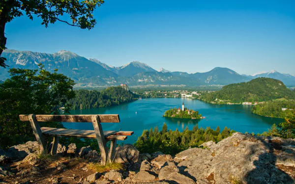 A serene landscape featuring a wooden bench overlooking Lake Bled, with the Assumption of Mary Church on an island, surrounded by mountains and clear blue skies.