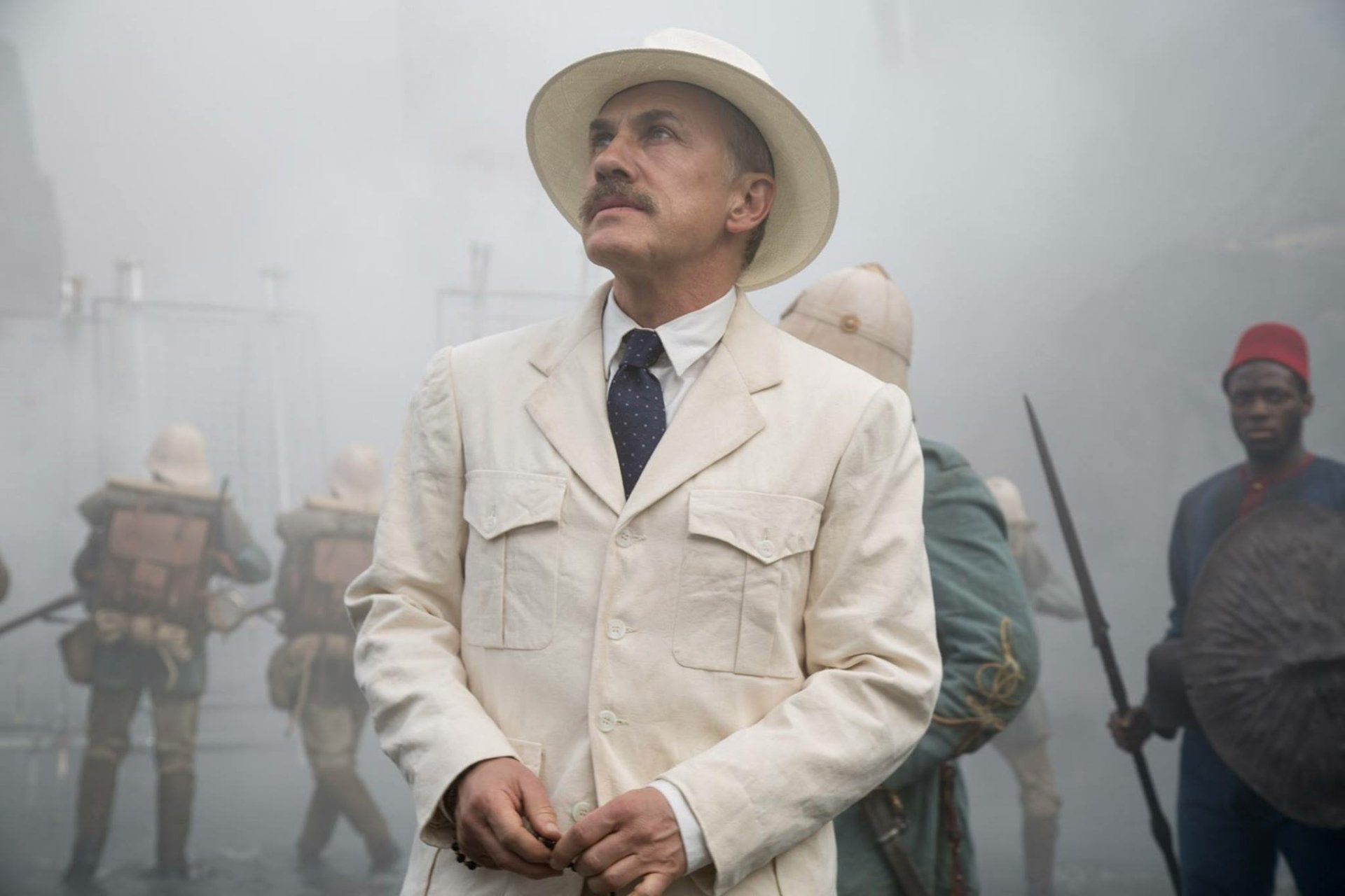 HD PC desktop wallpaper from the movie The Legend of Tarzan — a man in a white colonial suit and pith helmet stands amid a misty jungle camp with porters and armed men behind him.